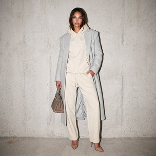 A model stands against a textured concrete wall wearing a light gray long coat over a cream hoodie and matching cream pants, holding a taupe handbag.