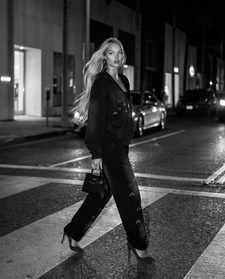 A woman walks across a city crosswalk at night wearing a black embroidered tracksuit and holding a small black handbag.