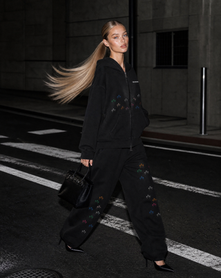 A woman walking on a city street at night wearing a black zip-up hoodie and matching black wide-leg pants, both with small multicolored embroidered motifs. She is carrying a black handbag.