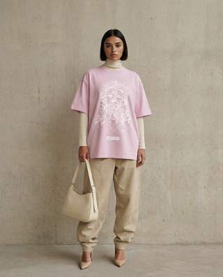 Person wearing a pink oversized t-shirt with a graphic design, beige pants, and holding a beige handbag against a plain background.