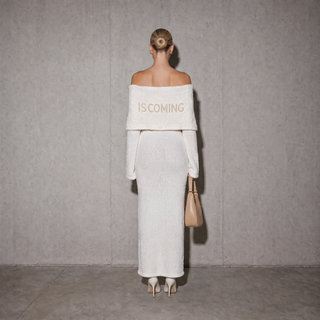 A model stands with her back to the camera wearing a textured off-the-shoulder white dress with 'IS COMING' printed in beige across the upper back. She is holding a beige handbag against a textured gray concrete wall.