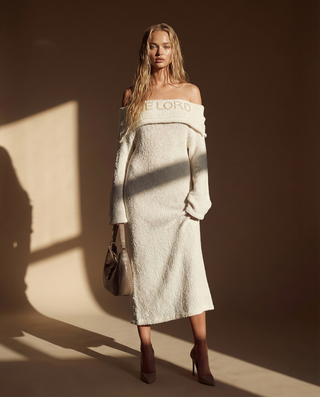 A woman models an off-the-shoulder cream bouclé midi dress with 'E LORD' embroidered across the neckline, styled with a taupe handbag and brown heels against a warm brown background with dramatic sunlight and shadows.