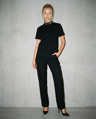 A woman modeling a black short-sleeve top and matching black trousers, standing against a textured gray concrete wall. The top features a mock neck with white text reading 'THE LORD'.