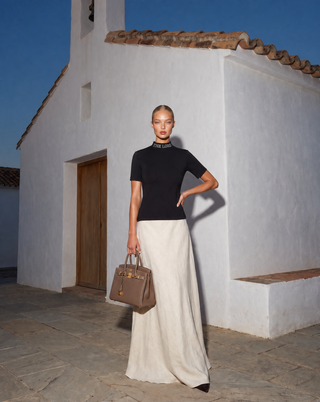 A model stands outdoors in front of a white stucco building, wearing a black short-sleeve top with a textured choker, a long cream skirt, and holding a brown leather handbag.