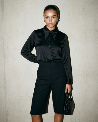 Woman wearing a black satin shirt and black shorts against a plain background