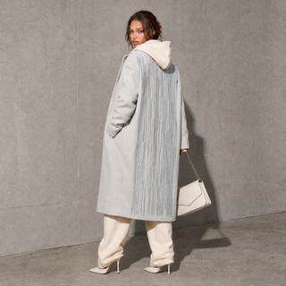 A woman stands against a textured gray wall wearing a long light gray hooded coat with vertical pleated detailing on the back, cream pants, and holding a white handbag.