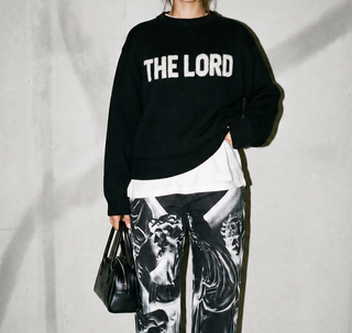 Person wearing a black sweater with 'THE LORD' text, holding a black handbag against a plain background